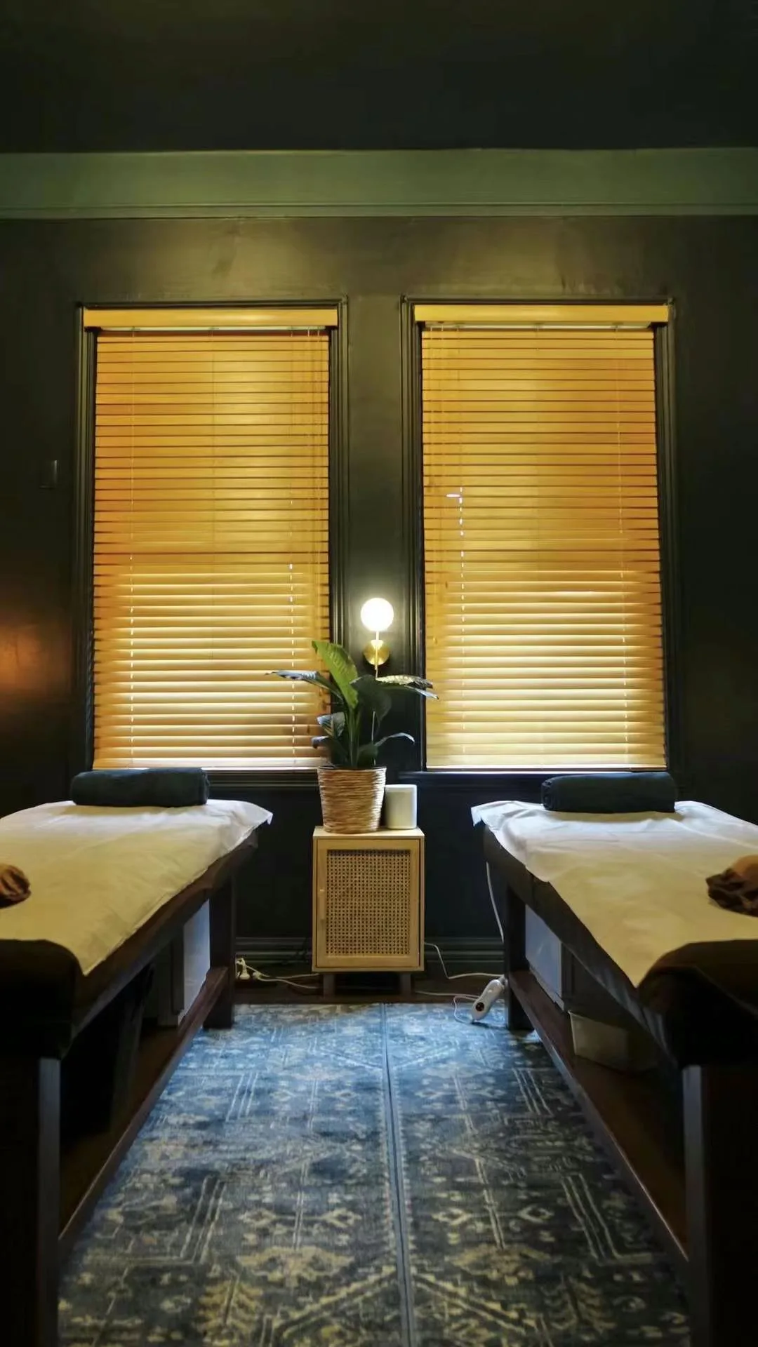 Twin-bed treatment room, amber venetian blinds glowing warm, a central aisle receding to tall windows