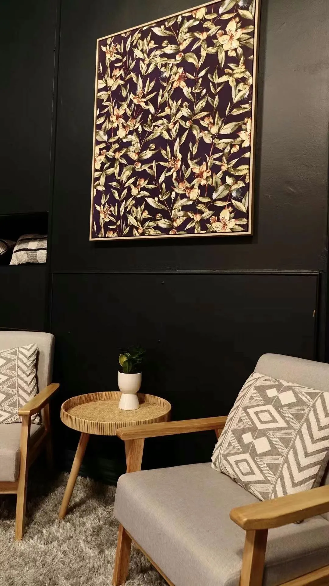 Dark lounge corner: large framed botanical canvas over two timber-armed chairs and a rattan side table