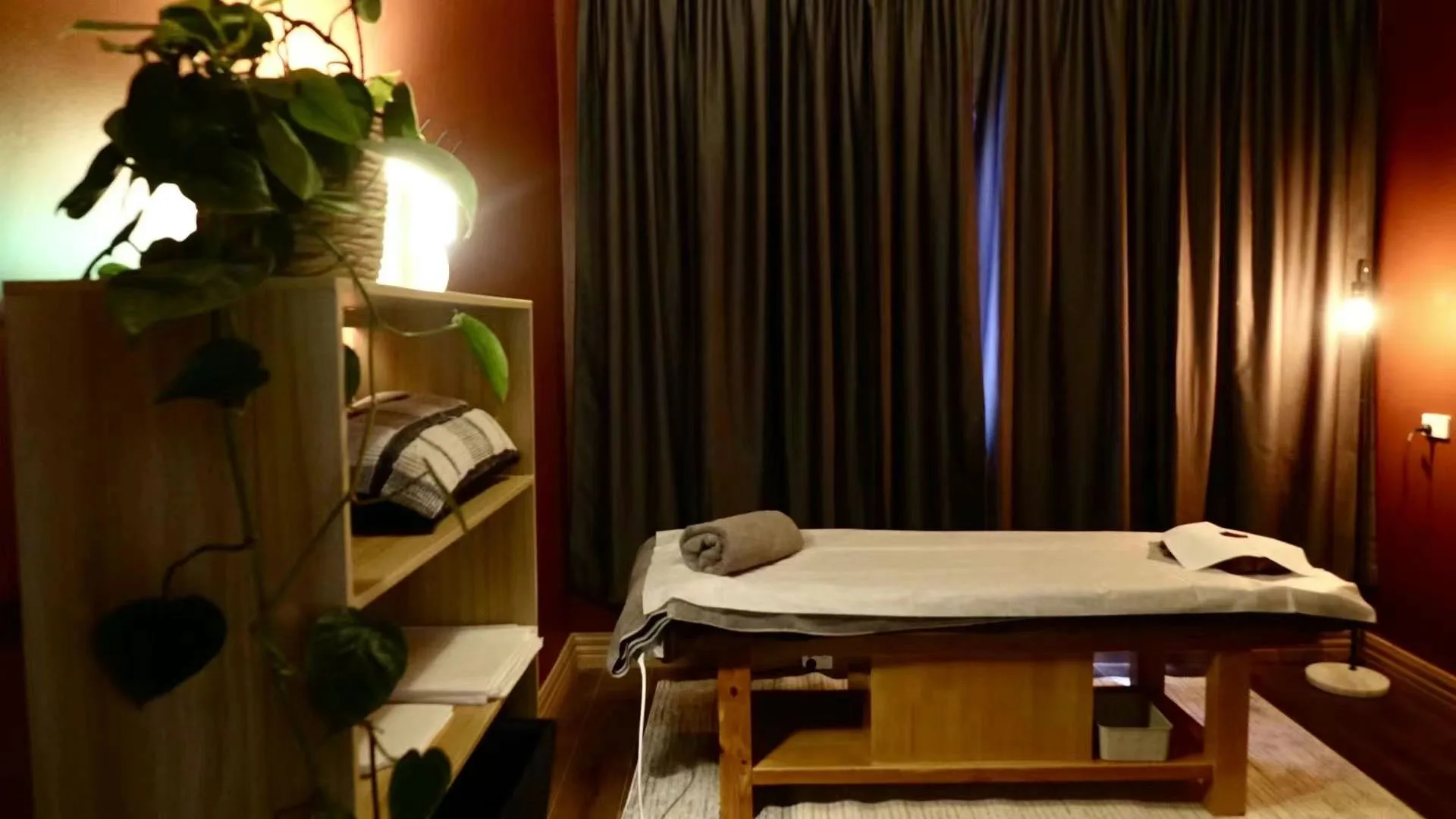 Single treatment room: dressed massage table with rolled towel, timber shelving and trailing pothos